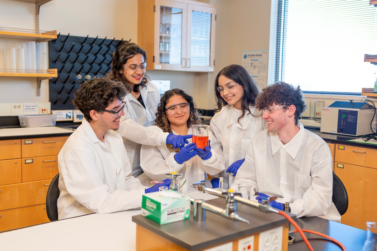 A group of five students grouped together in lab coats looking at a liquid