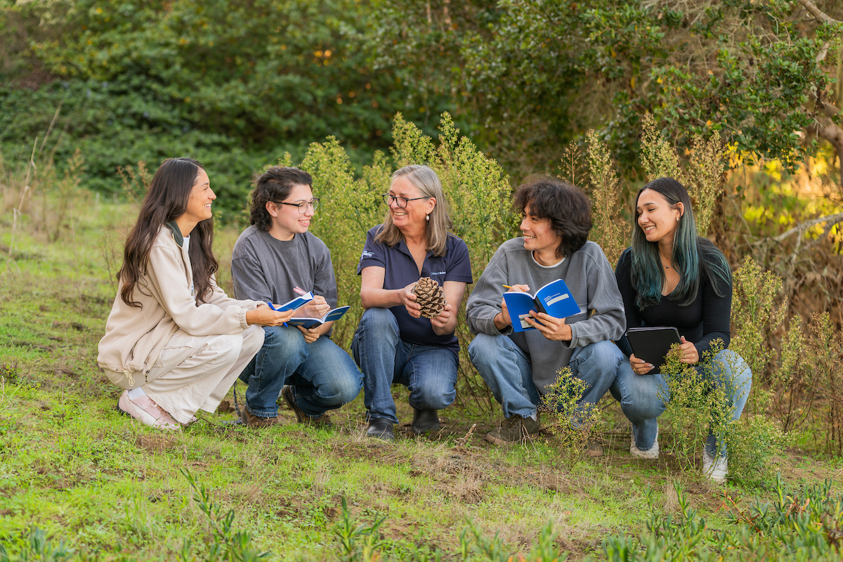 A group of four students and a professor standing outside in nature looking at each other