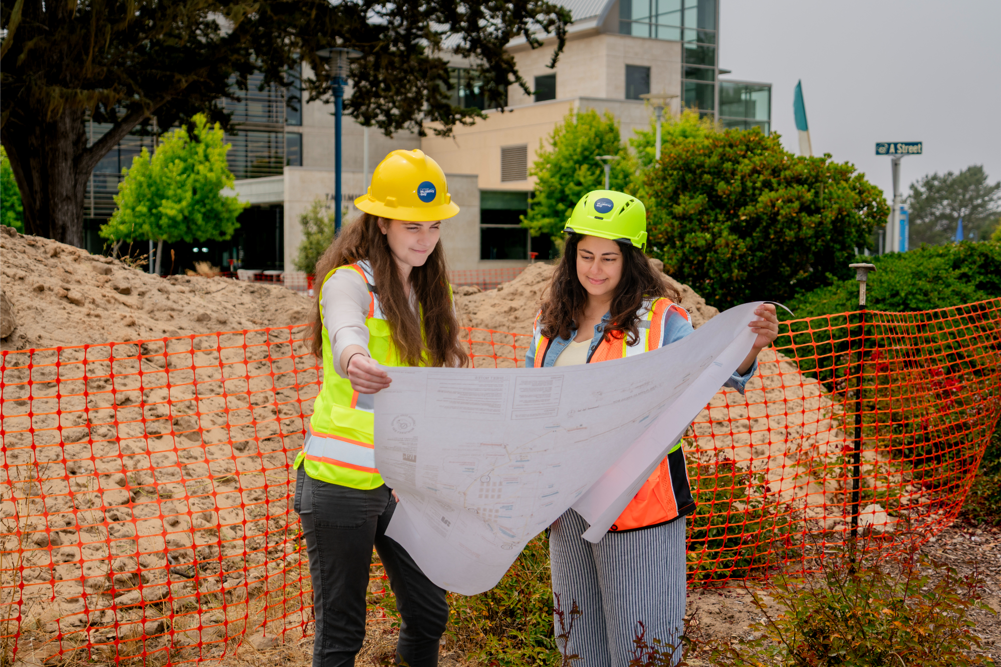 Two female construction professionals examine detailed building plans at a CSUMB construction site, with the library visible in the background.