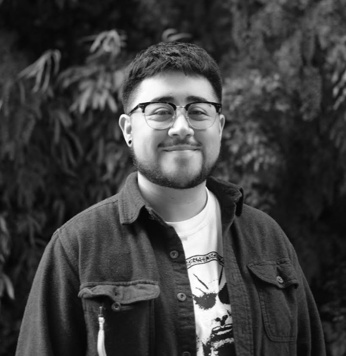 A person with short hair and glasses is smiling outdoors, wearing a printed T-shirt and a button-up shirt, with foliage in the background.