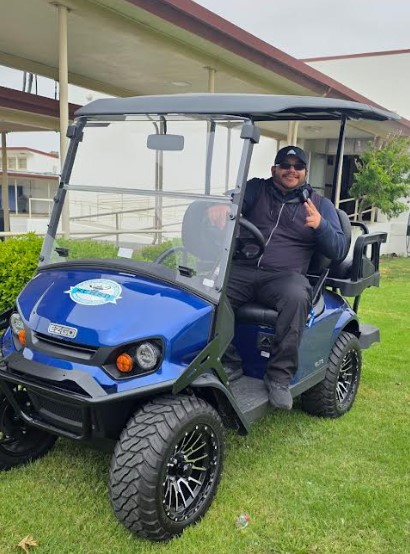 A person in dark clothing and sunglasses is sitting in a blue golf cart on a grassy area, smiling and flashing a peace sign.