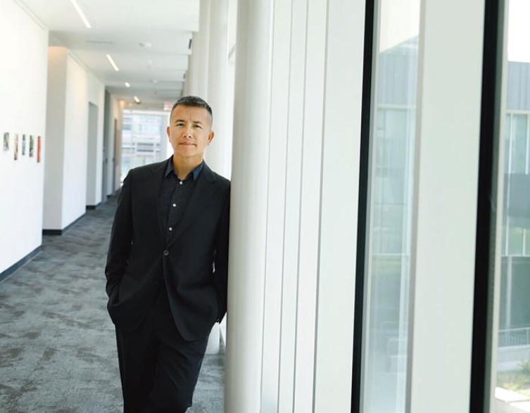 A man in a dark suit stands in a bright hallway, leaning casually against a white column with his hands in his pockets. Natural light streams in from large windows beside him.