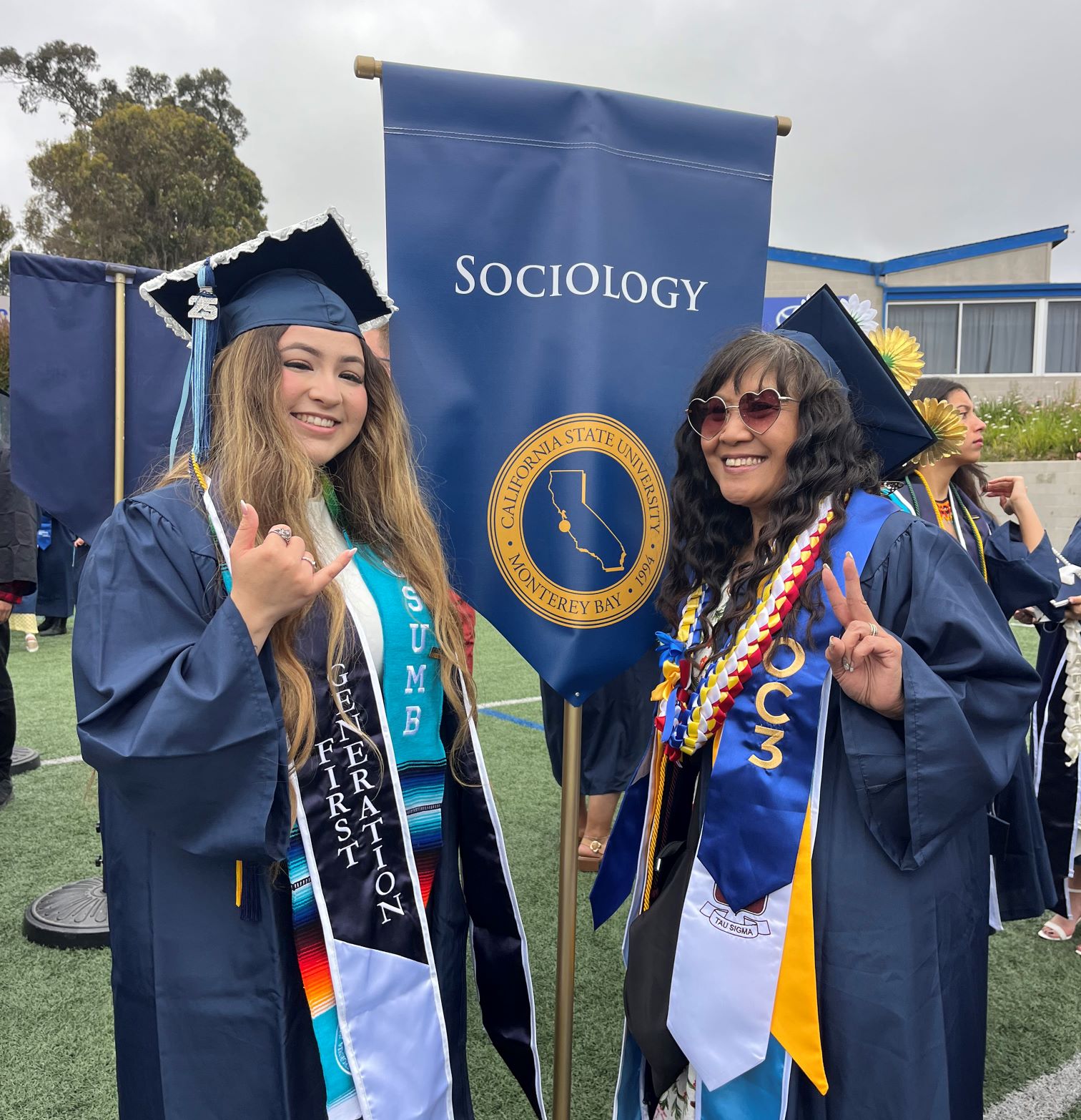Two female sociology graduates in their caps and gowns standing in front of the Sociology banner at the commencement ceremony