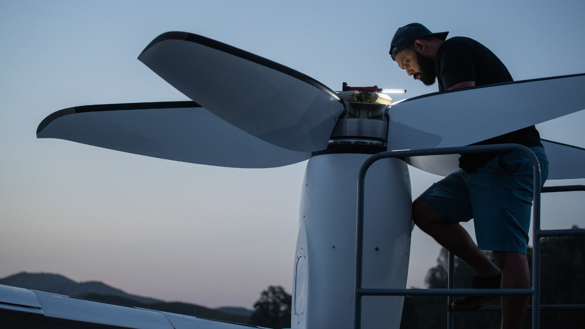 A worker performs maintenance on a large drone or aircraft propeller at dusk, standing on a platform and illuminated by a small light, with hills silhouetted in the background.