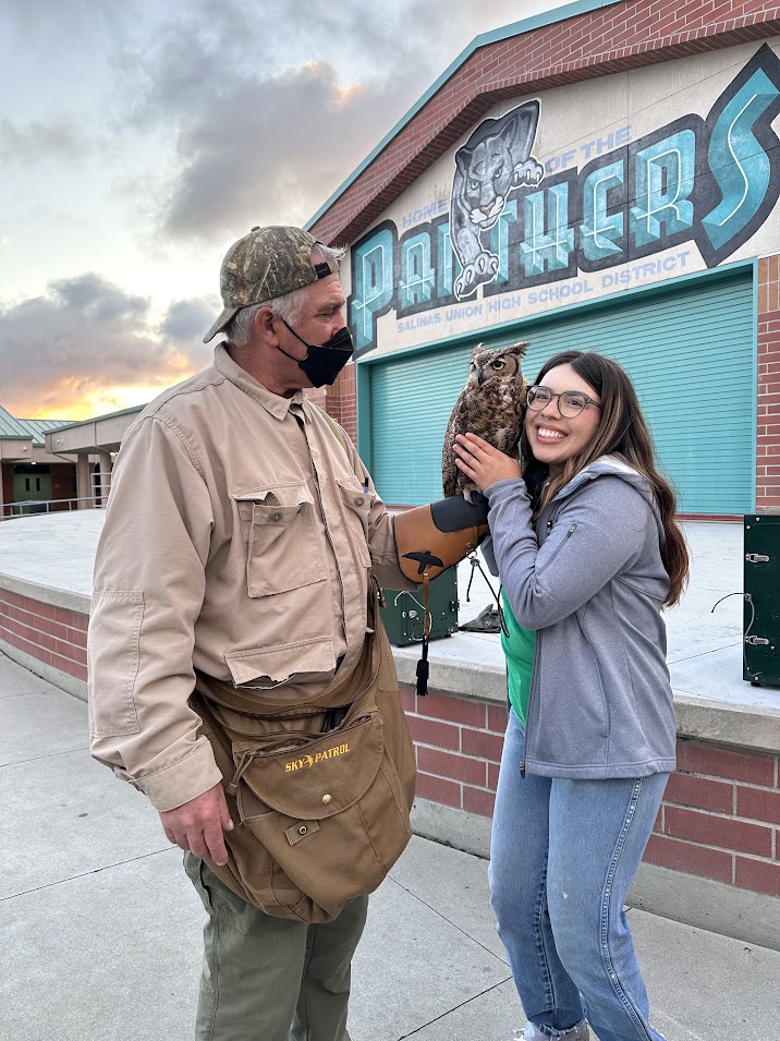 Cecelia smiling and holding an owl; the owl is perched on its handler's arm. The handler is dressed in his uniform.