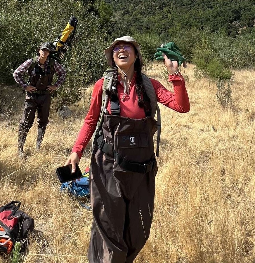 Destiny smiling and doing a funny pose for the camera while standing in a grassy area. There is a person with his hands on his hips while looking at her in an exasperated manner