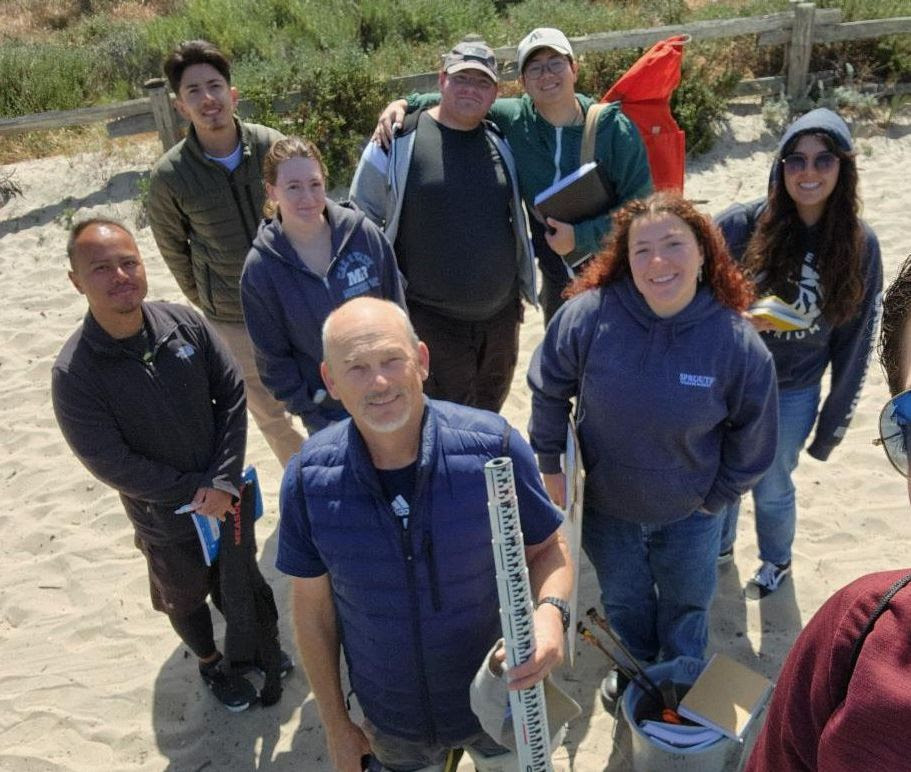 Doug Smith smiling while holding a piece of equipment in his hands. He stands near a bank of a dry creek