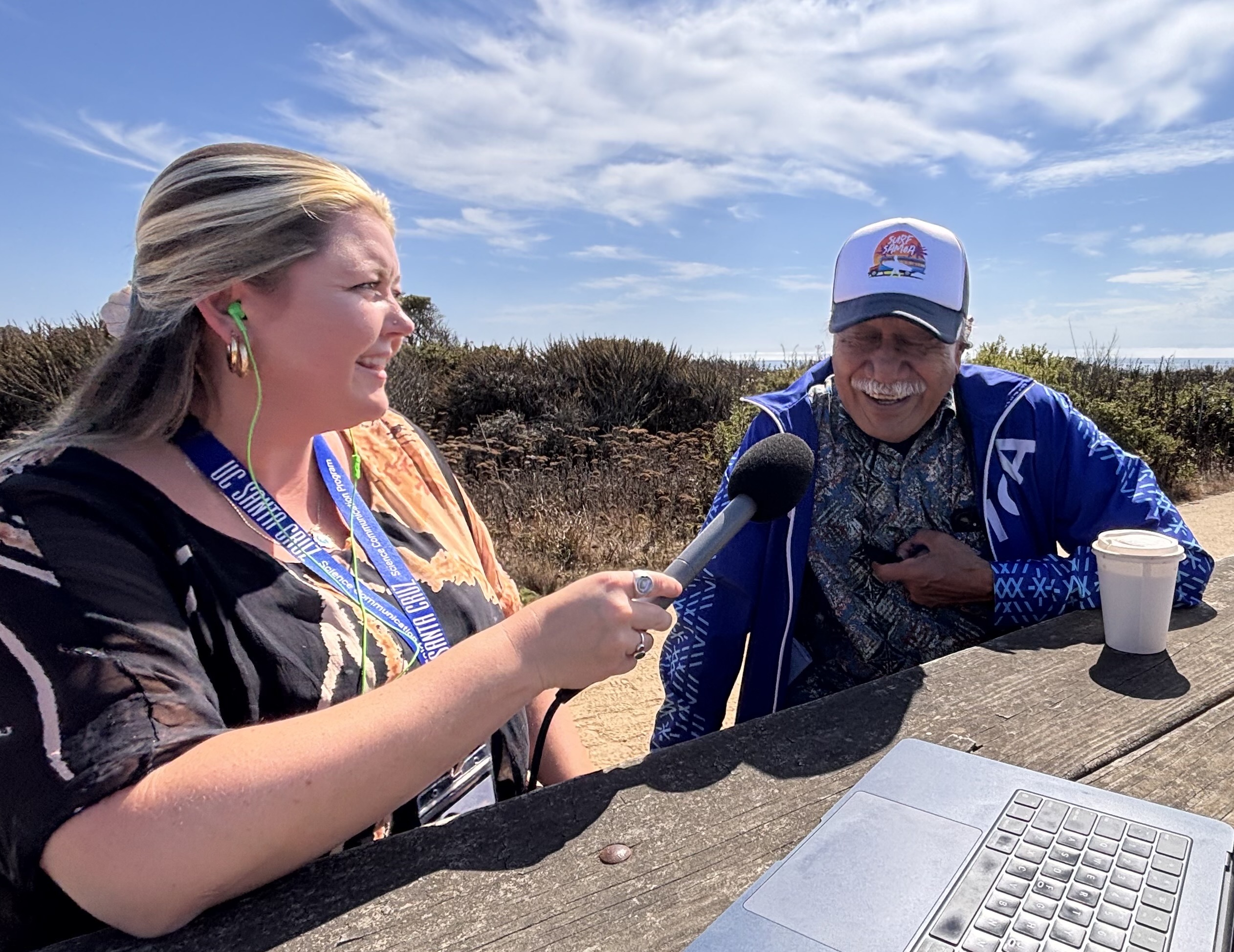 Katie is sitting and talking with an indigenous Samoan chief. Both are sitting at a table, Katie is facing away from the camera. She holds a microphone in her hand