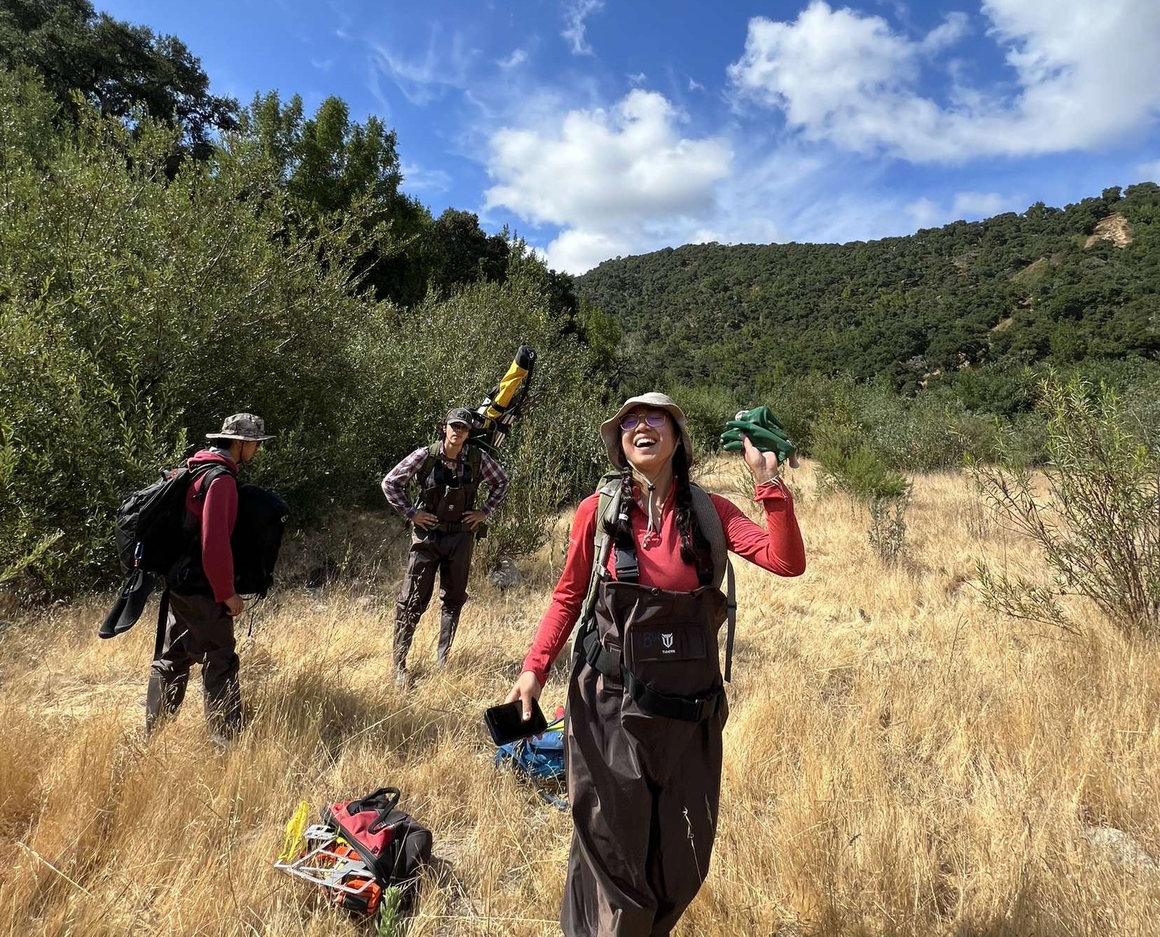 Three people in waders stand in a grassy field with outdoor gear, under a blue sky with clouds.