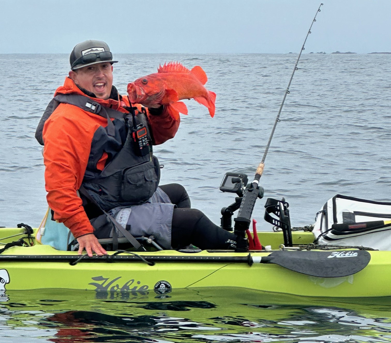 Person in a green kayak holding a large orange fish with fishing gear on a calm water body.