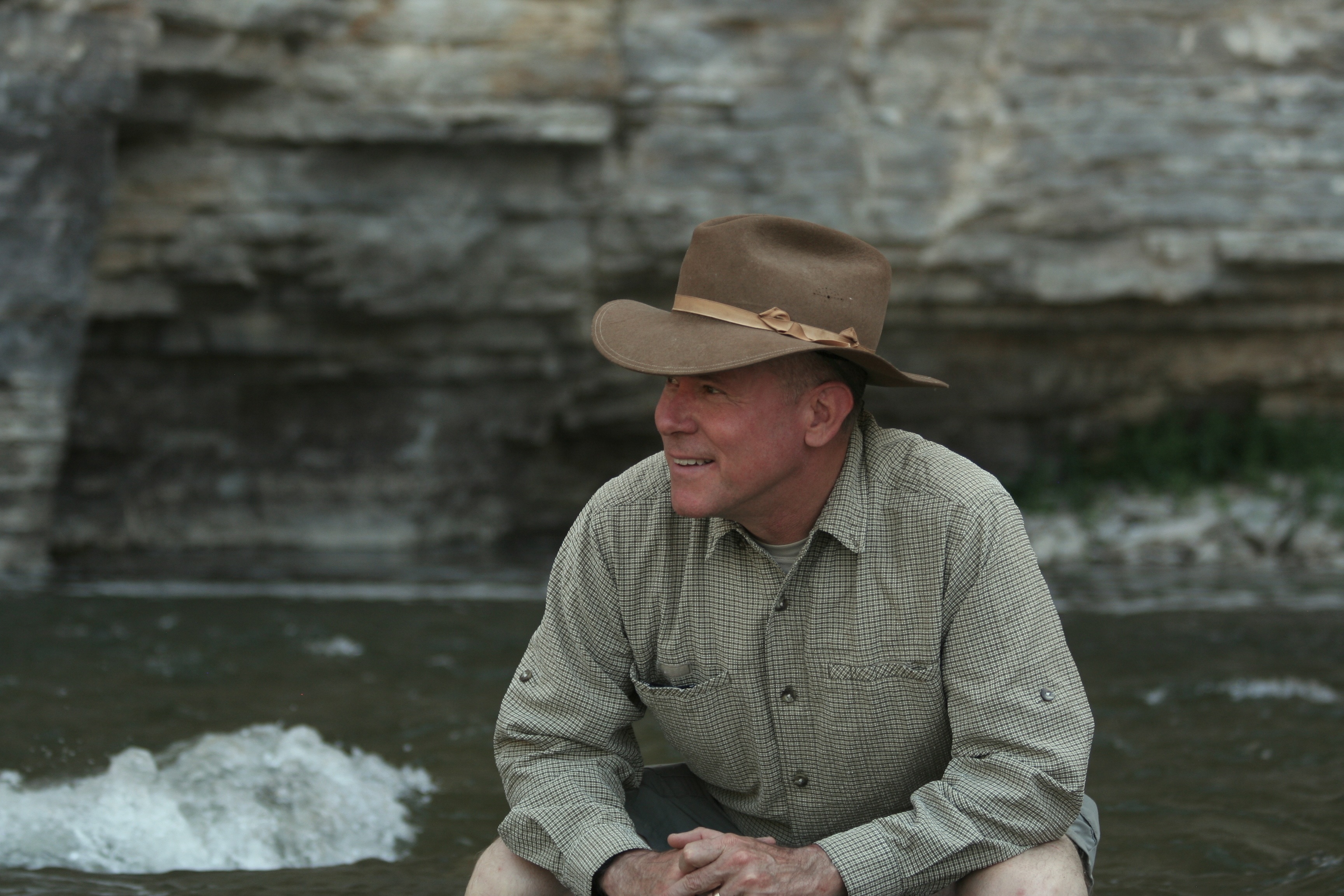 John wearing a brown hat sitting in front of a rock and river