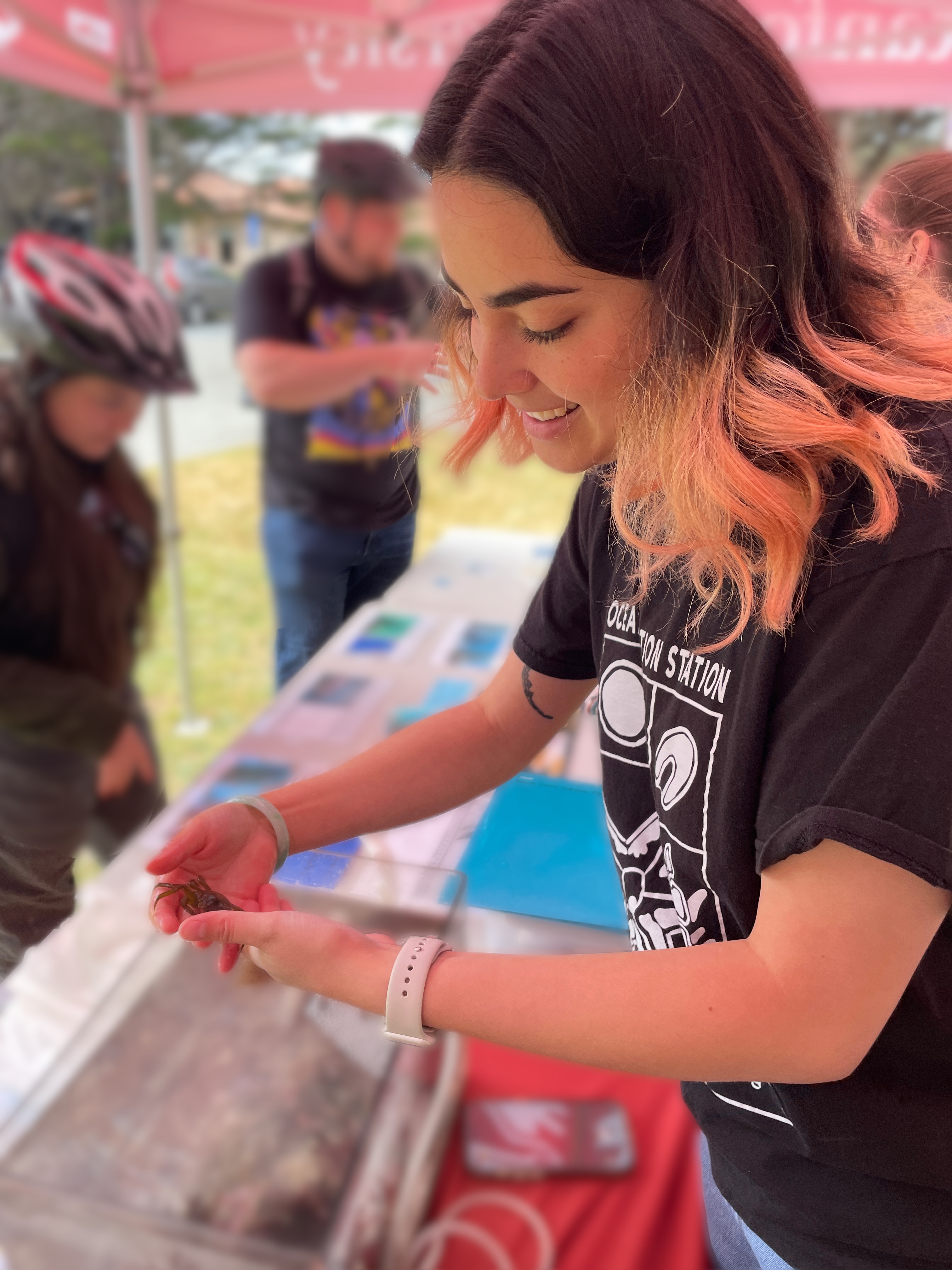 A woman holds a small crab at an outdoor event under a tent.