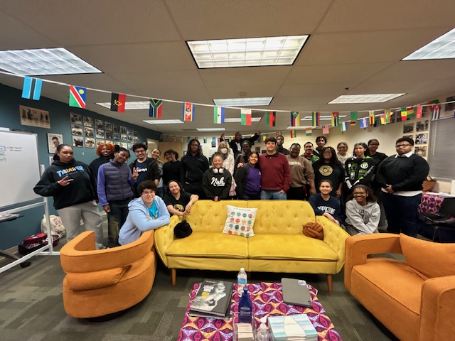 A large group of smiling students standing around a yellow couch and orange chairs