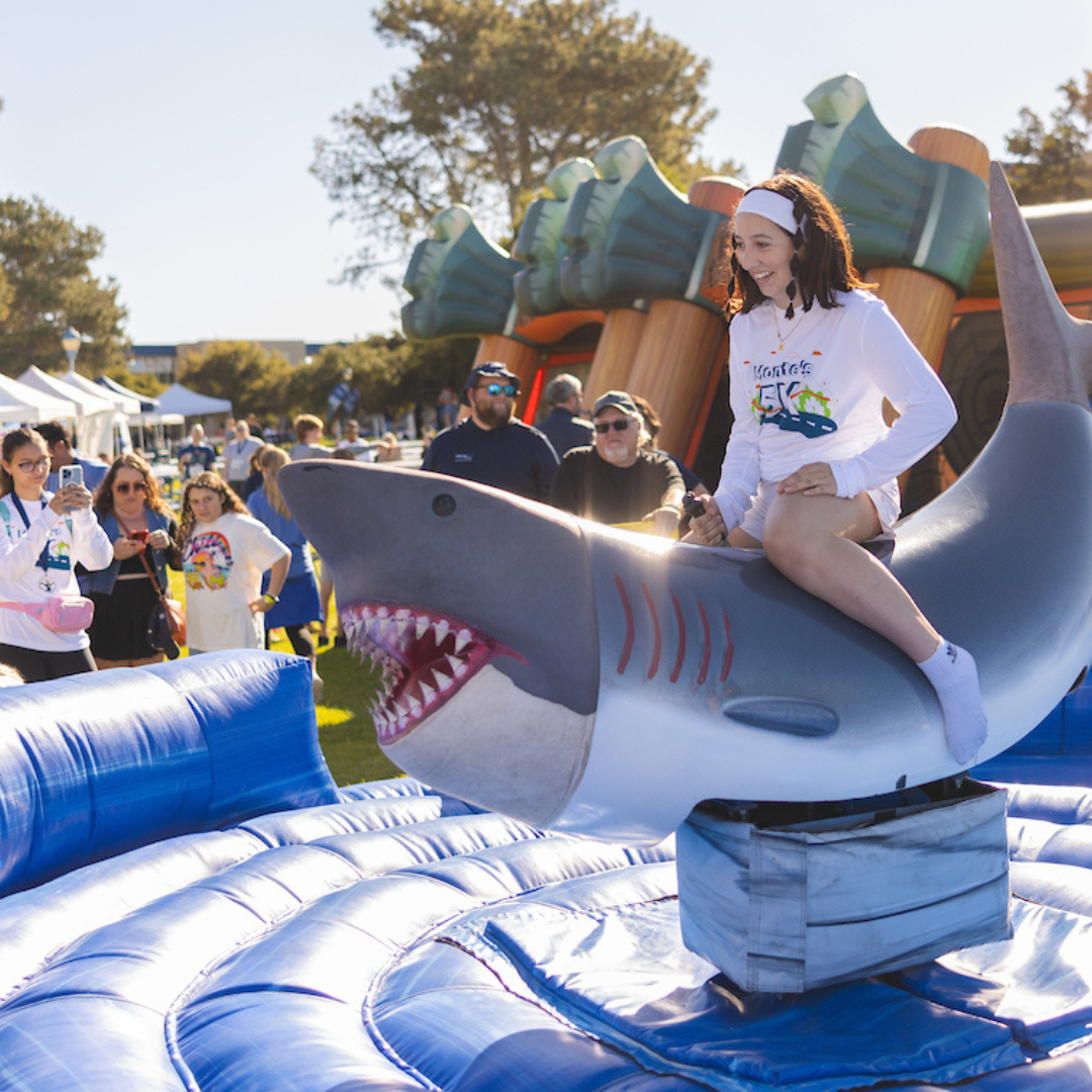 Student riding mechanical shark and people playing carnival games in the background at MB Madness event