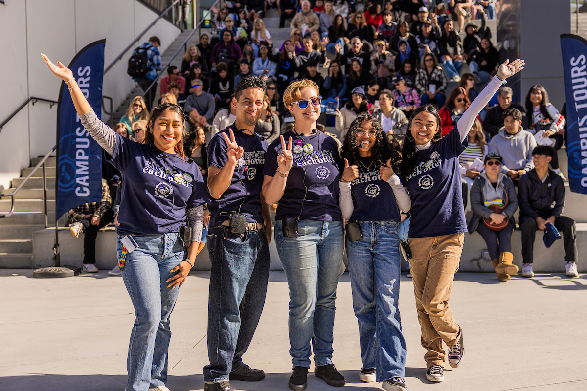 5 Orientation Leaders posing towards camera campus with tour group in the back