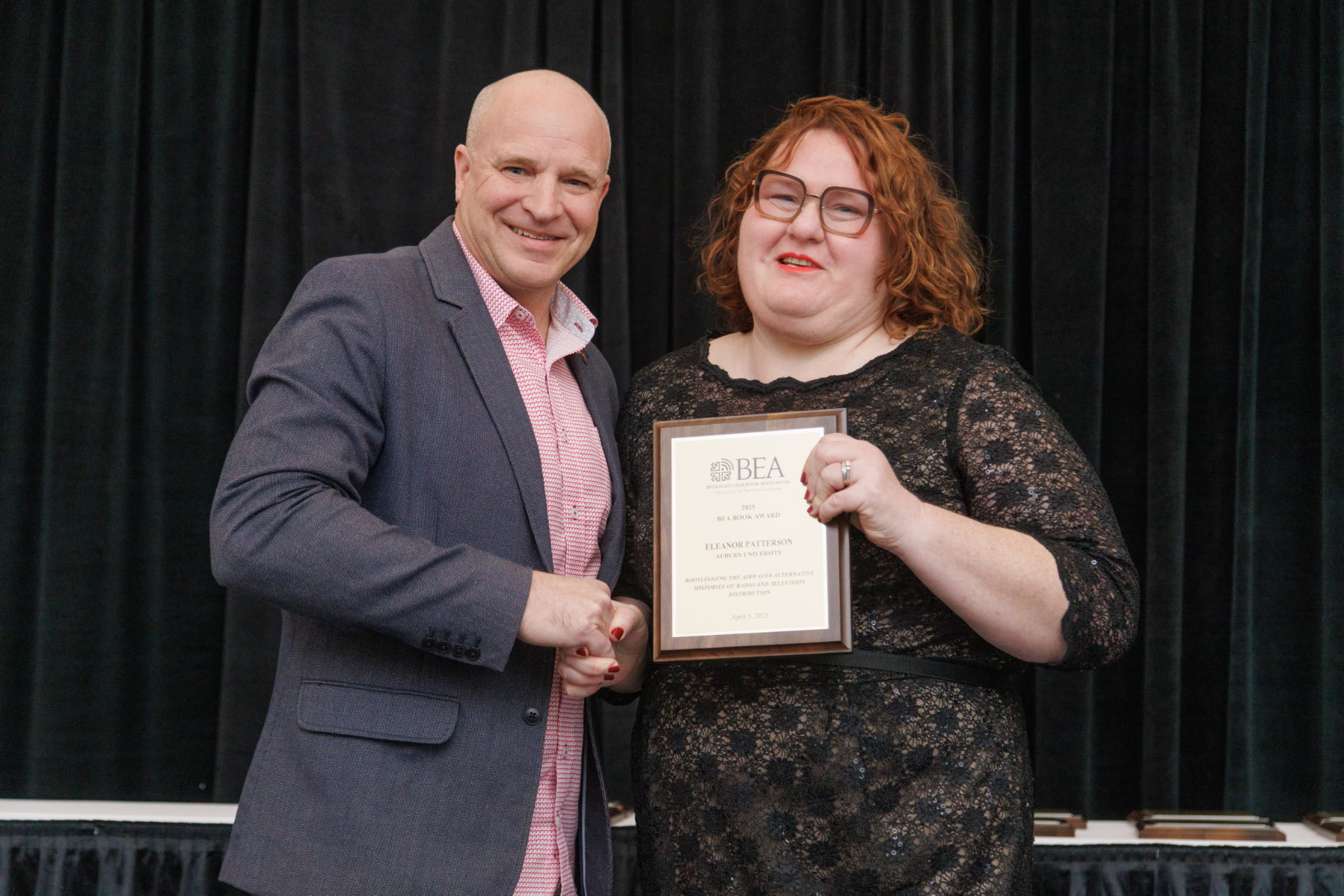 Two people are standing in front of a black curtain; one is holding a plaque that reads 
