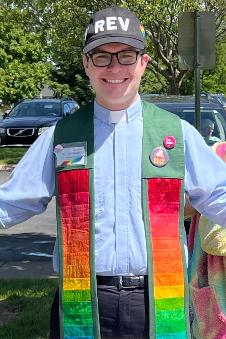 A smiling man wearing a clerical collar, rainbow stole, and a black cap that says 