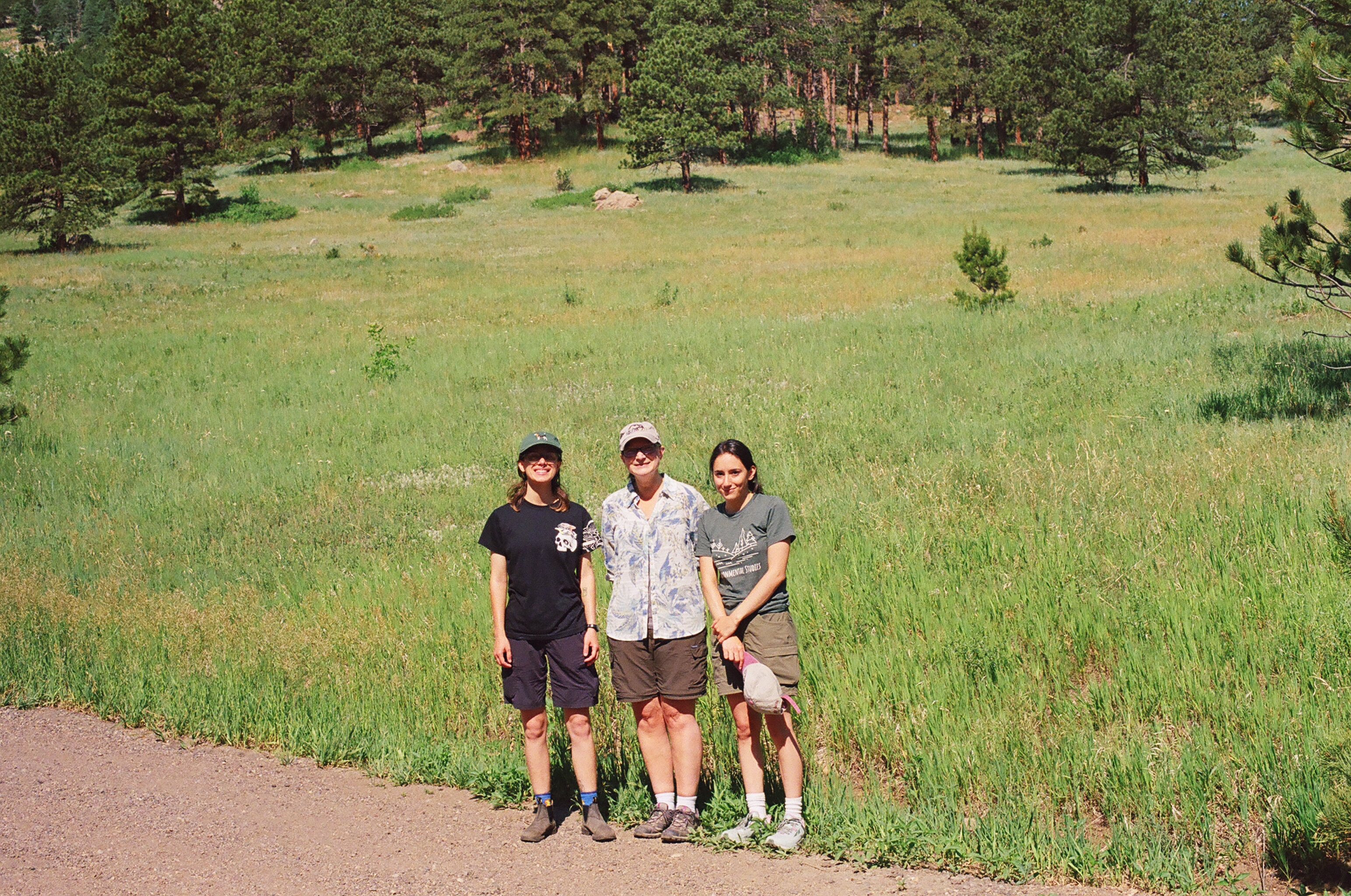 Three people standing on a dirt path in front of a grassy field and pine trees.