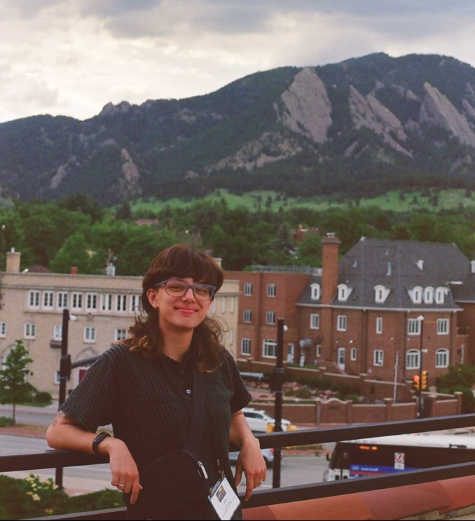 A person with glasses standing in front of buildings and green mountains.