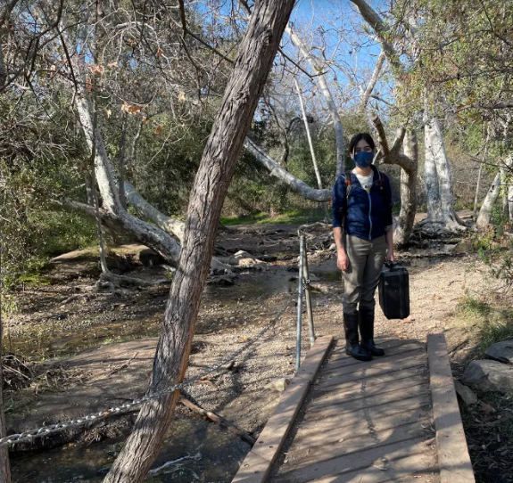 Connies stands with research equipment on a wooden bridge above a small stream