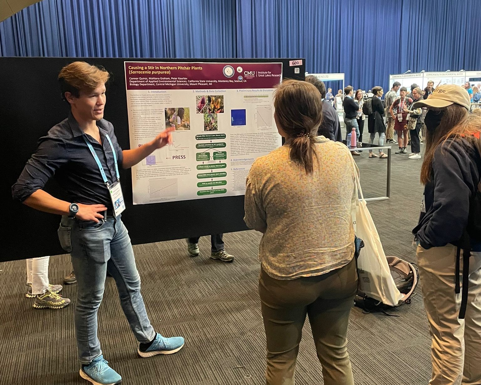 Conor pointing to his research poster while talking to a person. Conor's standing to the side with a hand on his hip.