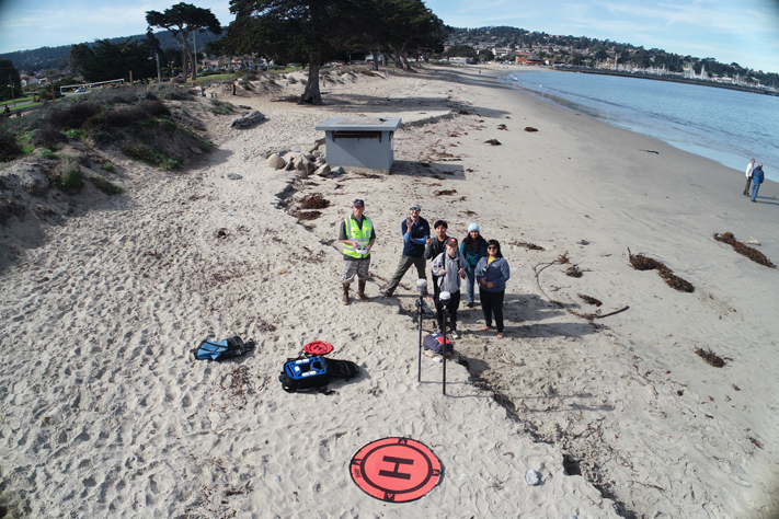 A group of students standing on a beach while a drone takes their picture