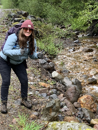 Emma Debasitis flashing a thumbs up to the camera while standing next to a creek/river