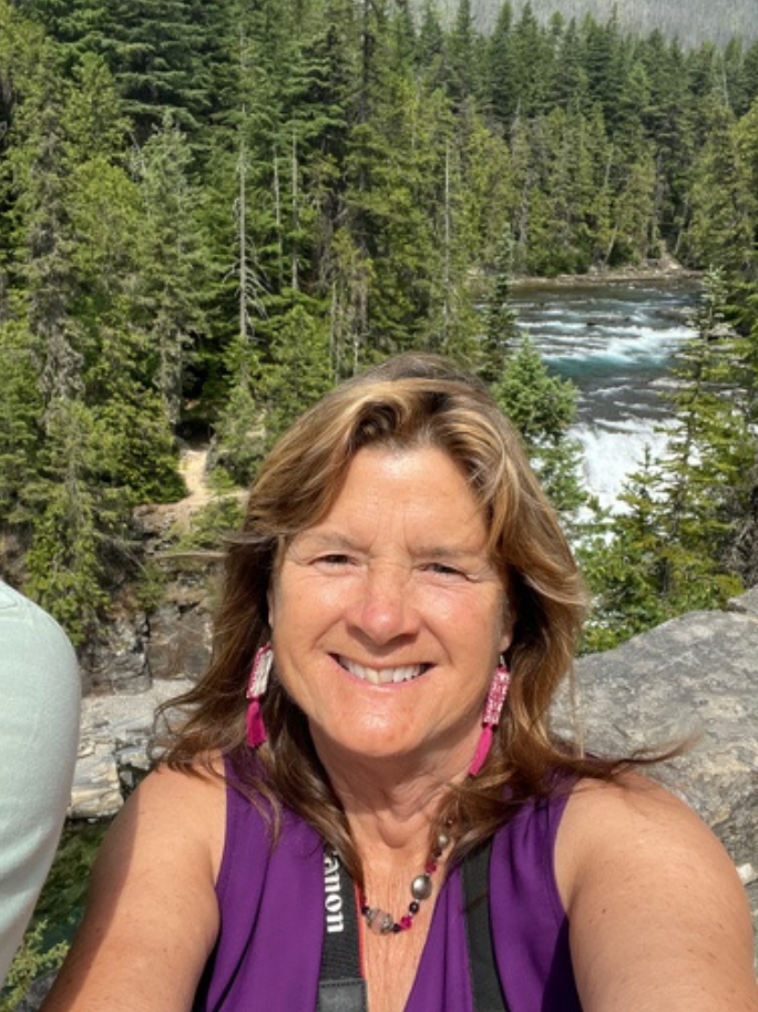 Woman taking a selfie in front of a dense forest and river with white water rapids.