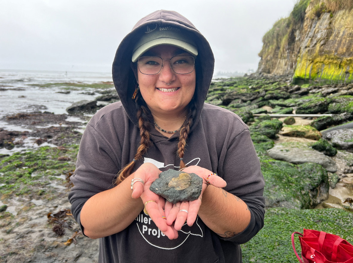 Kaitlyn holding a fossilized rock on a rocky, seaweed-covered coastline.