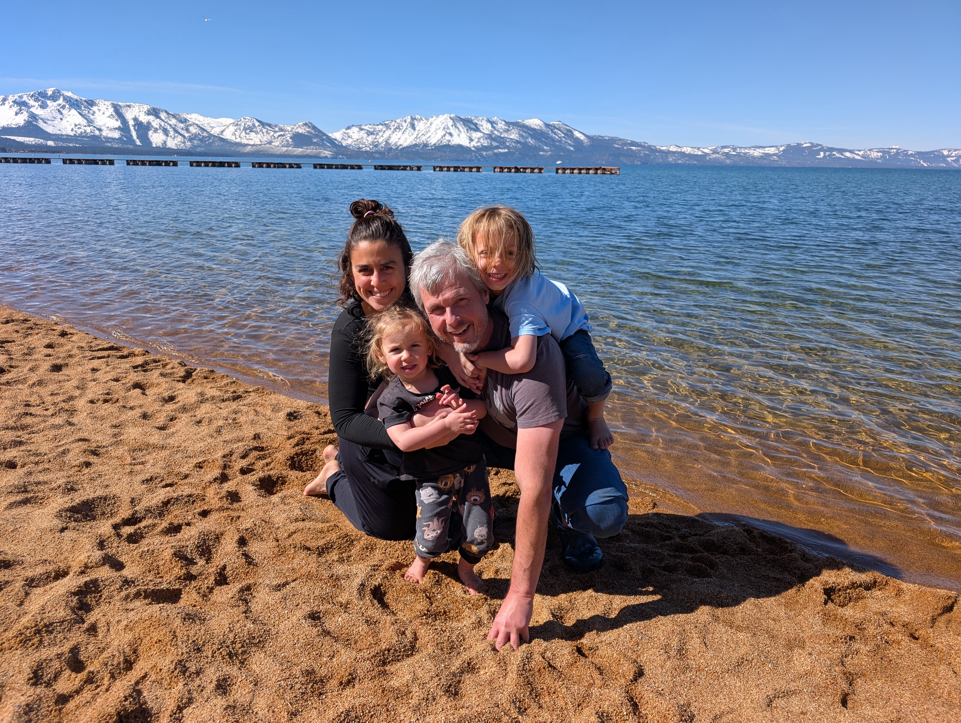 Kelly with his family at a lake with mountains in the background. Kely is holding a child while another child is on his back. Next to Kelly is his wife