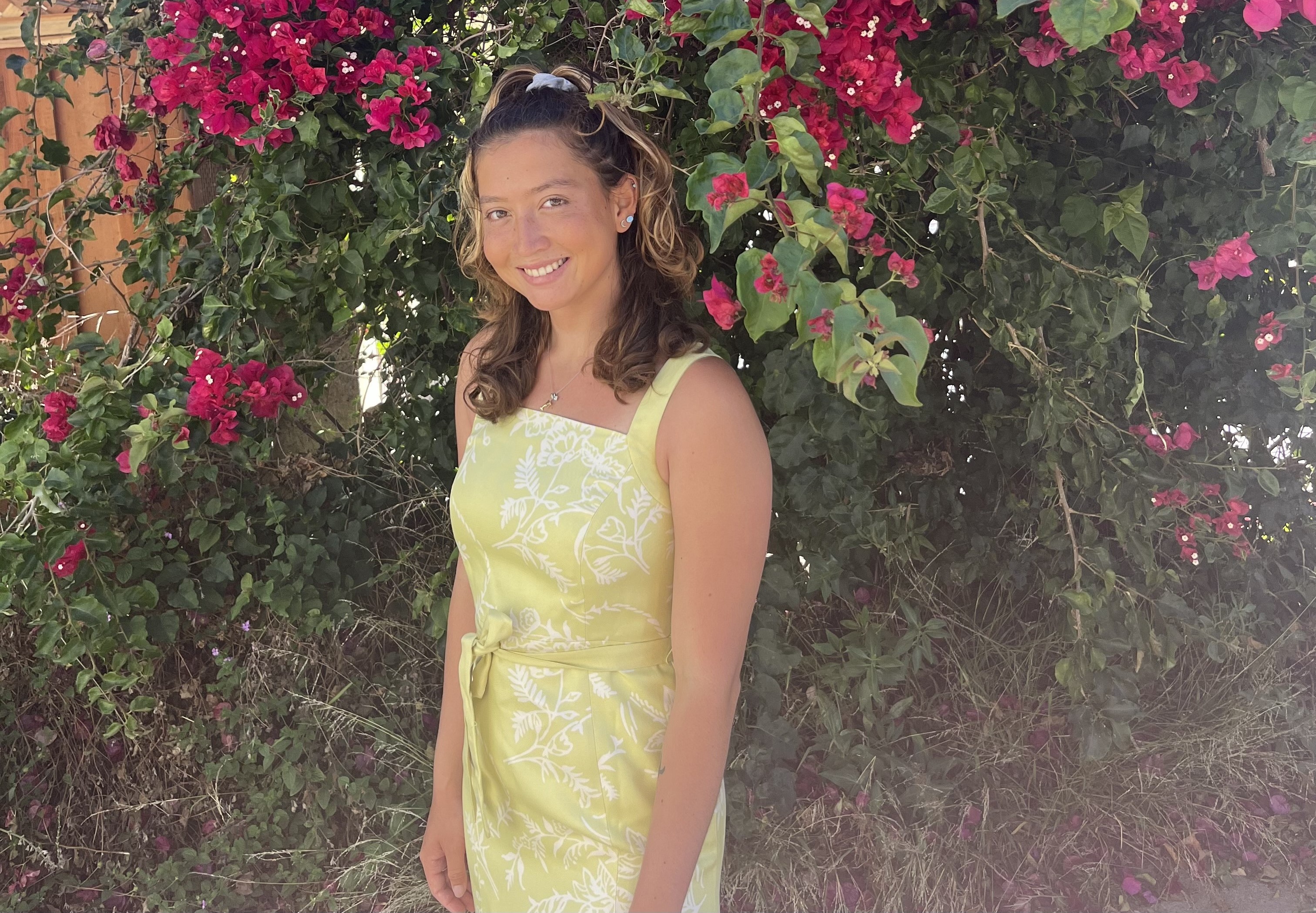 Alyssa smiling and standing in front of a tree with pink and white flowers. Alyssa is wearing a yellow dress with white plant stencils