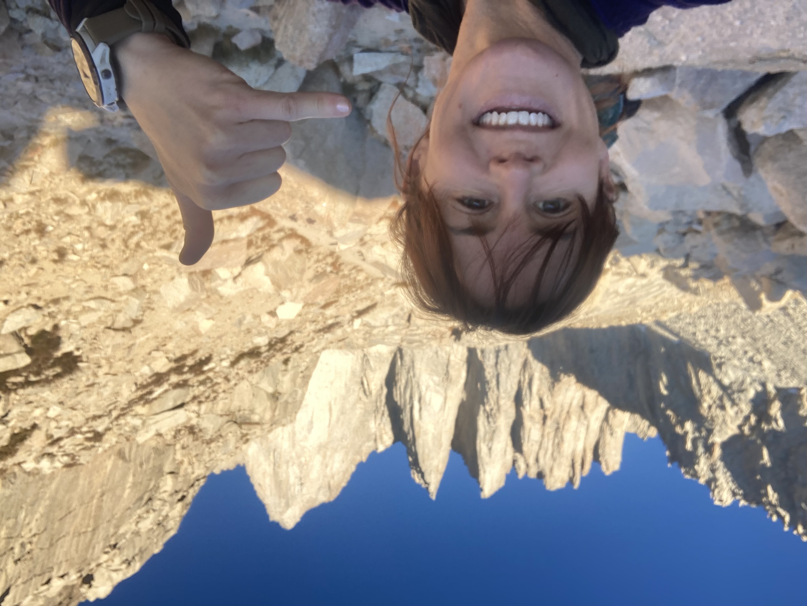 Woman smiling and making a shaka sign in front of a mountain range under a blue sky.