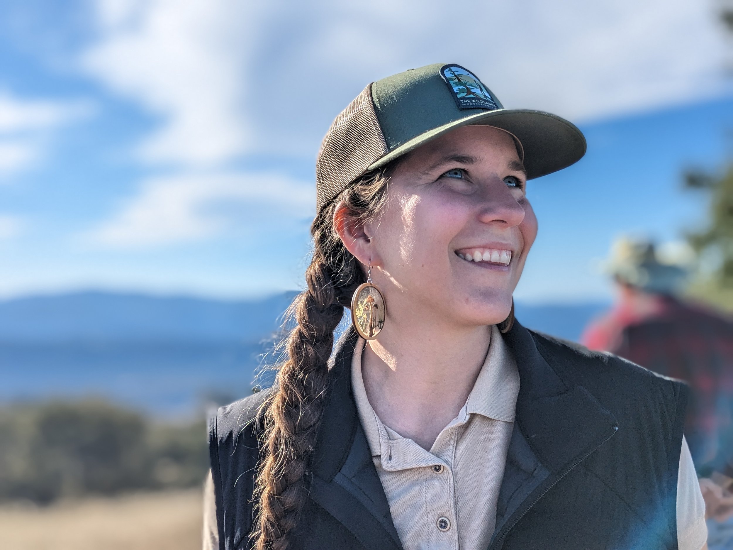 A person with a braid wearing a green hat and dark vest smiles outdoors with a mountainous landscape in the background.