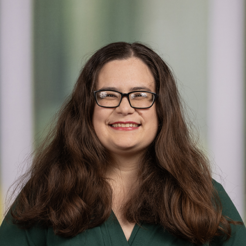 Kierra smiling while wearing a green shirt, against a soft, blurred background. She is wearing glasses and has long, brown hair