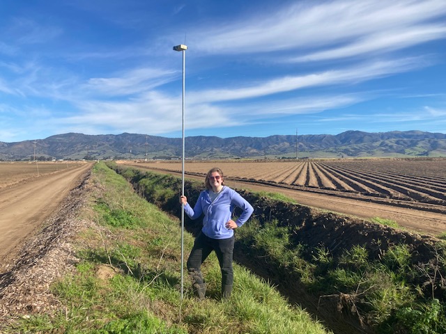 Person holding a tall GPS or surveying pole next to a dirt road and agricultural fields with mountains in the background.