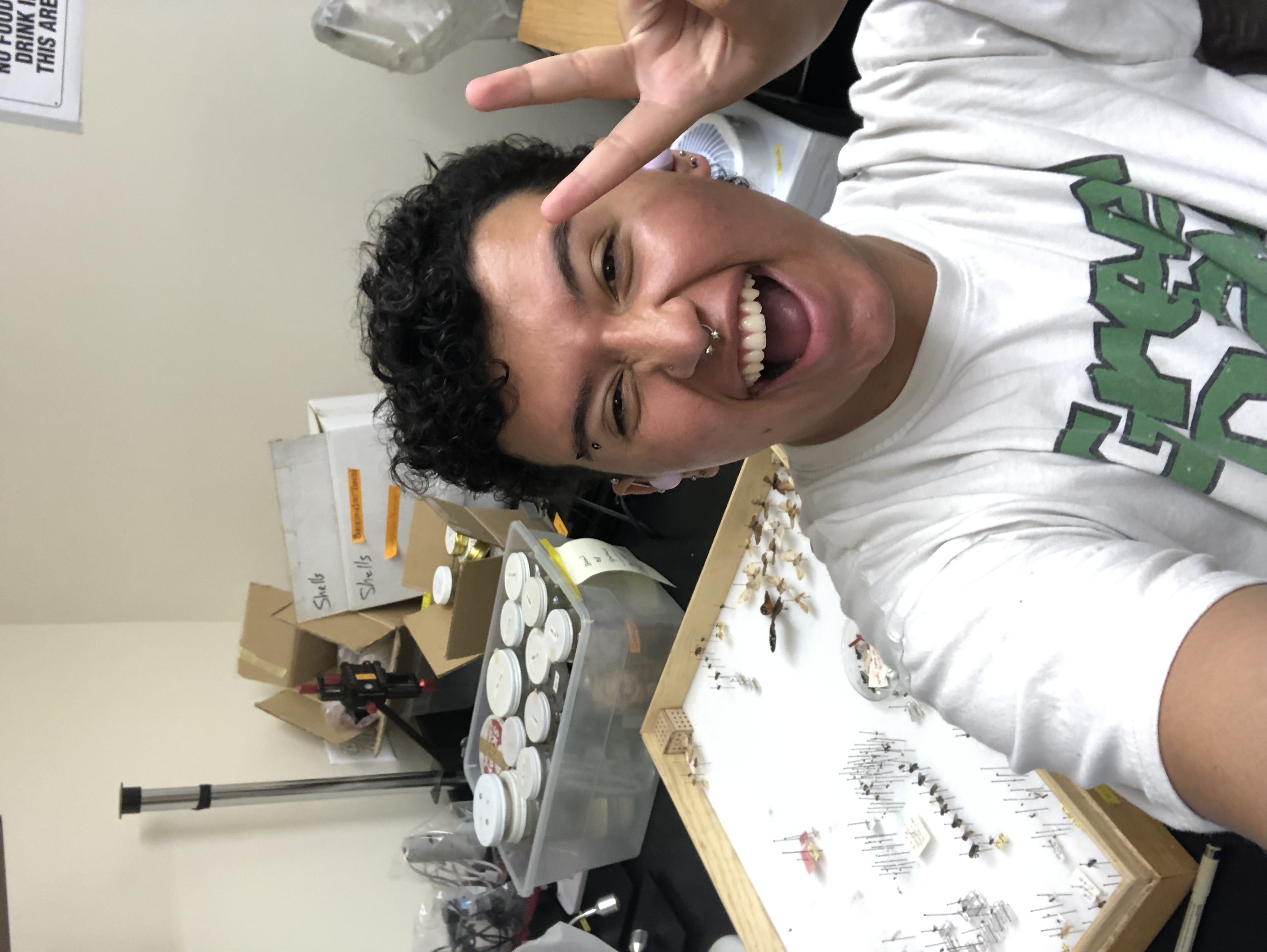Miles is taking a selfie and flashing a peace sign. Behind Miles is a wooden box containing pinned insect specimens. Next to that box is a container filled with jars and other pieces of scientific equipment