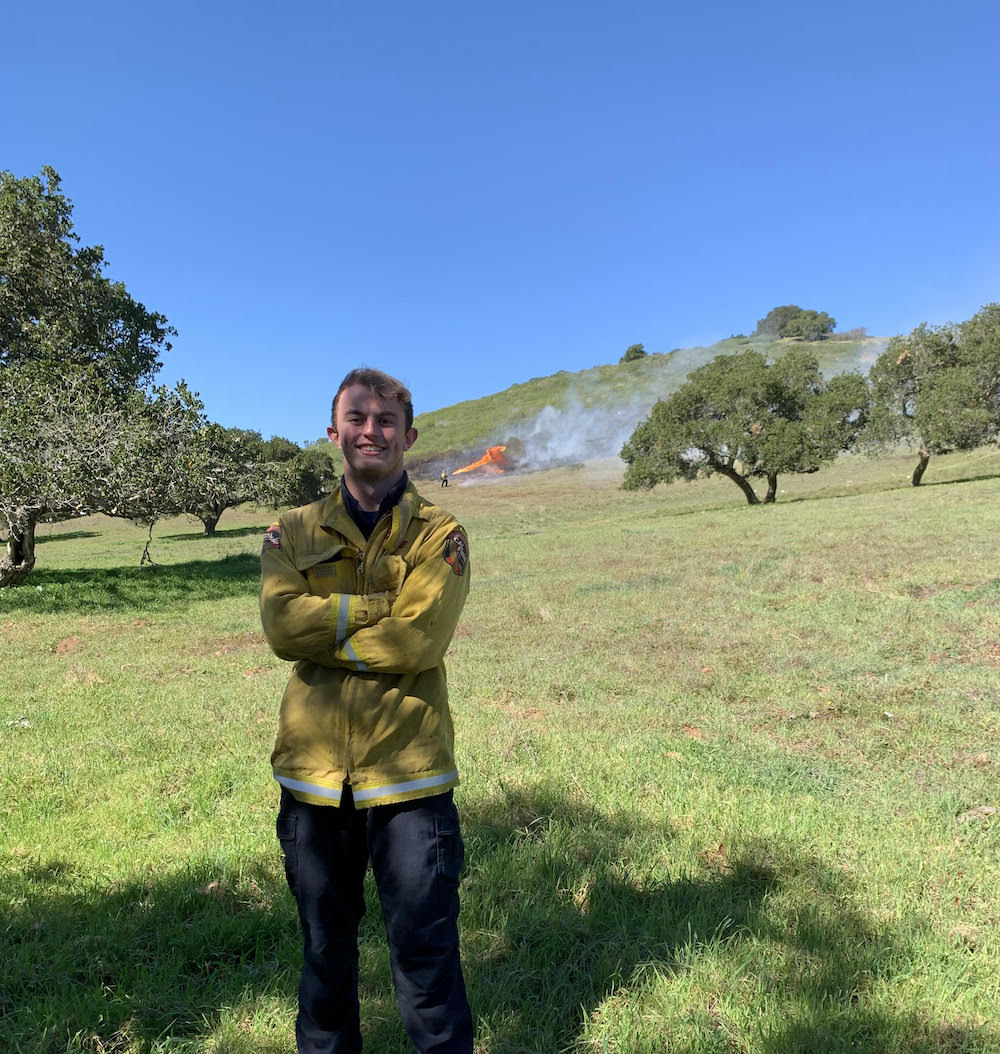 Sam poses in a field with his arms crossed in front of his chest, and a CAL Fire jacket on