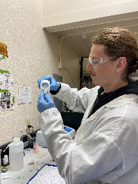 Justin, wearing a lab coat and safety goggles, pouring a liquid into a small glass container.