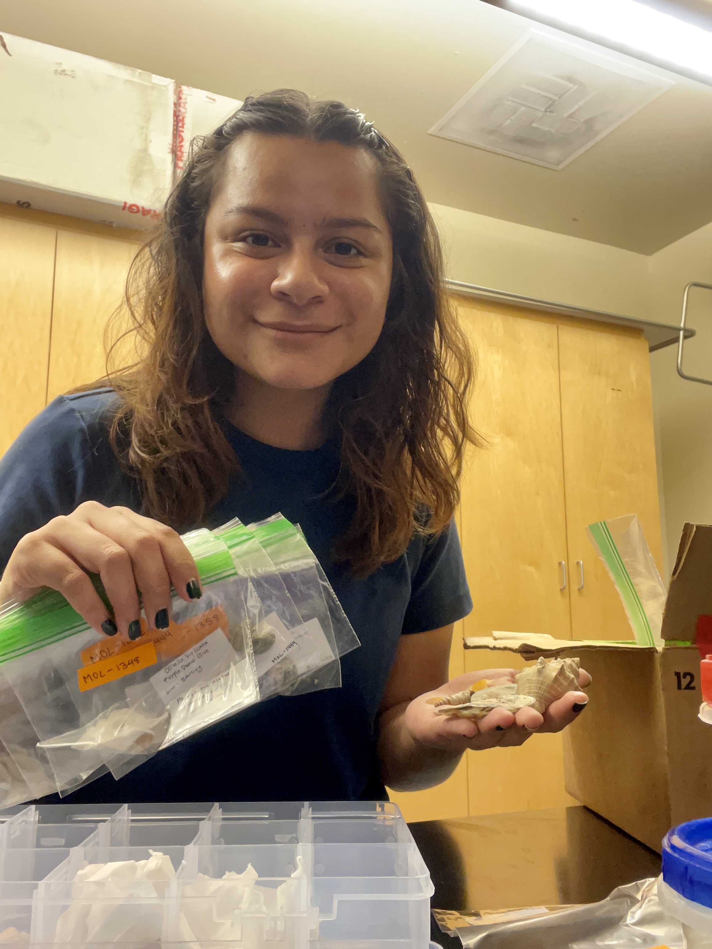 Emily is smiling for the picture while holding multiple sandwich bags filled with various shell specimens in one hand. The other hand is holding multiple shells