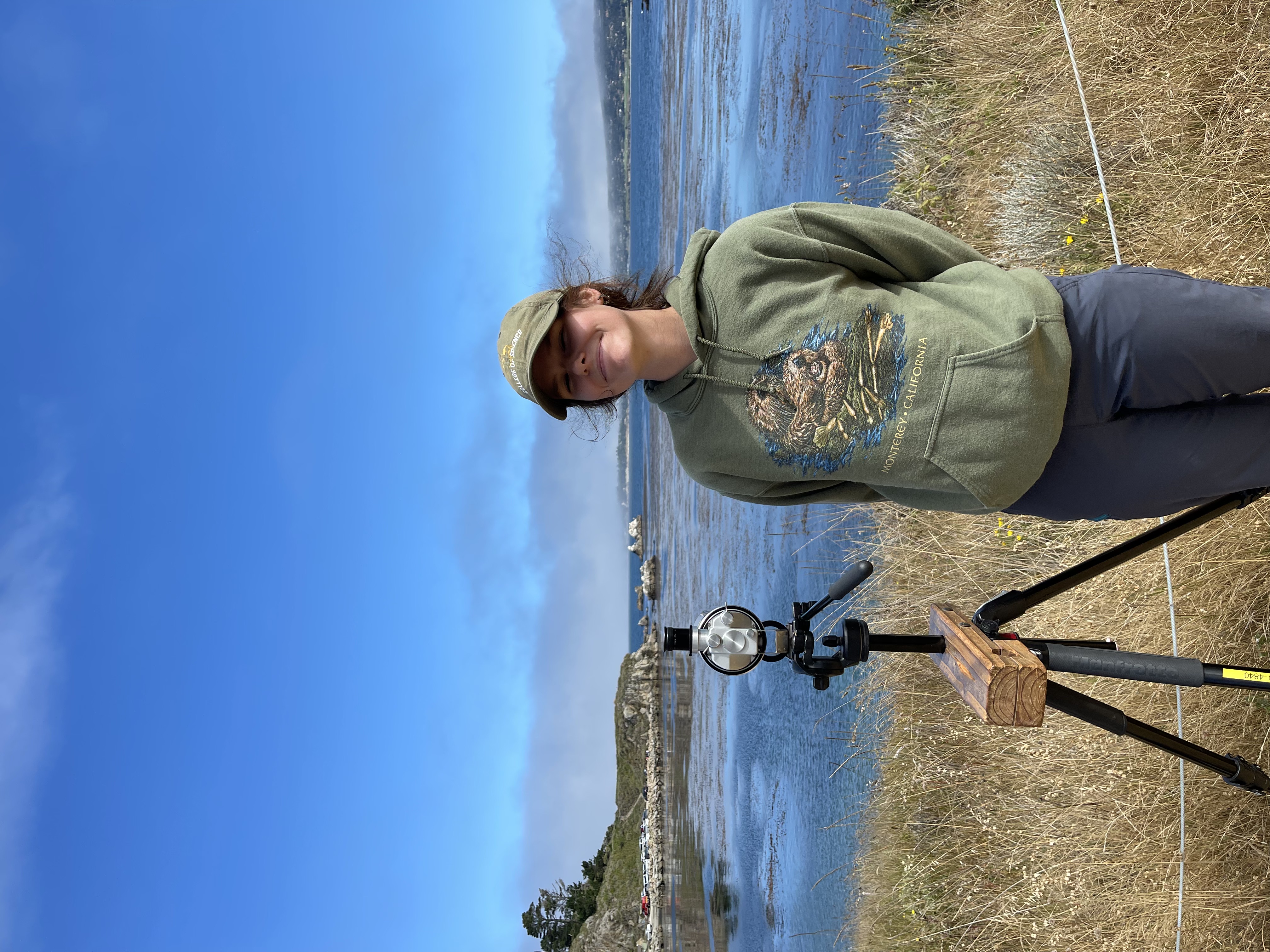 The image depicts a person standing next to a tripod-mounted instrument near a coastal area. The person is wearing a brownish-green baseball cap and a green hoodie with an illustration of sea otters and the words 