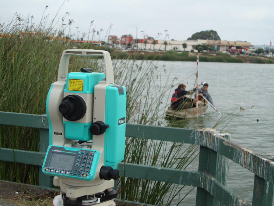 Nikon total station on a tripod by the water with three people in a rowboat in the background.