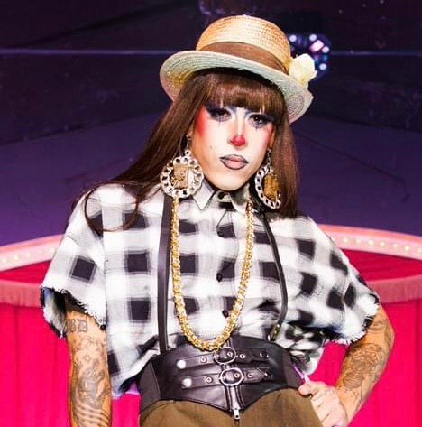 Headshot of Kochina Rude, a professional drag queen, wearing plaid and a hat