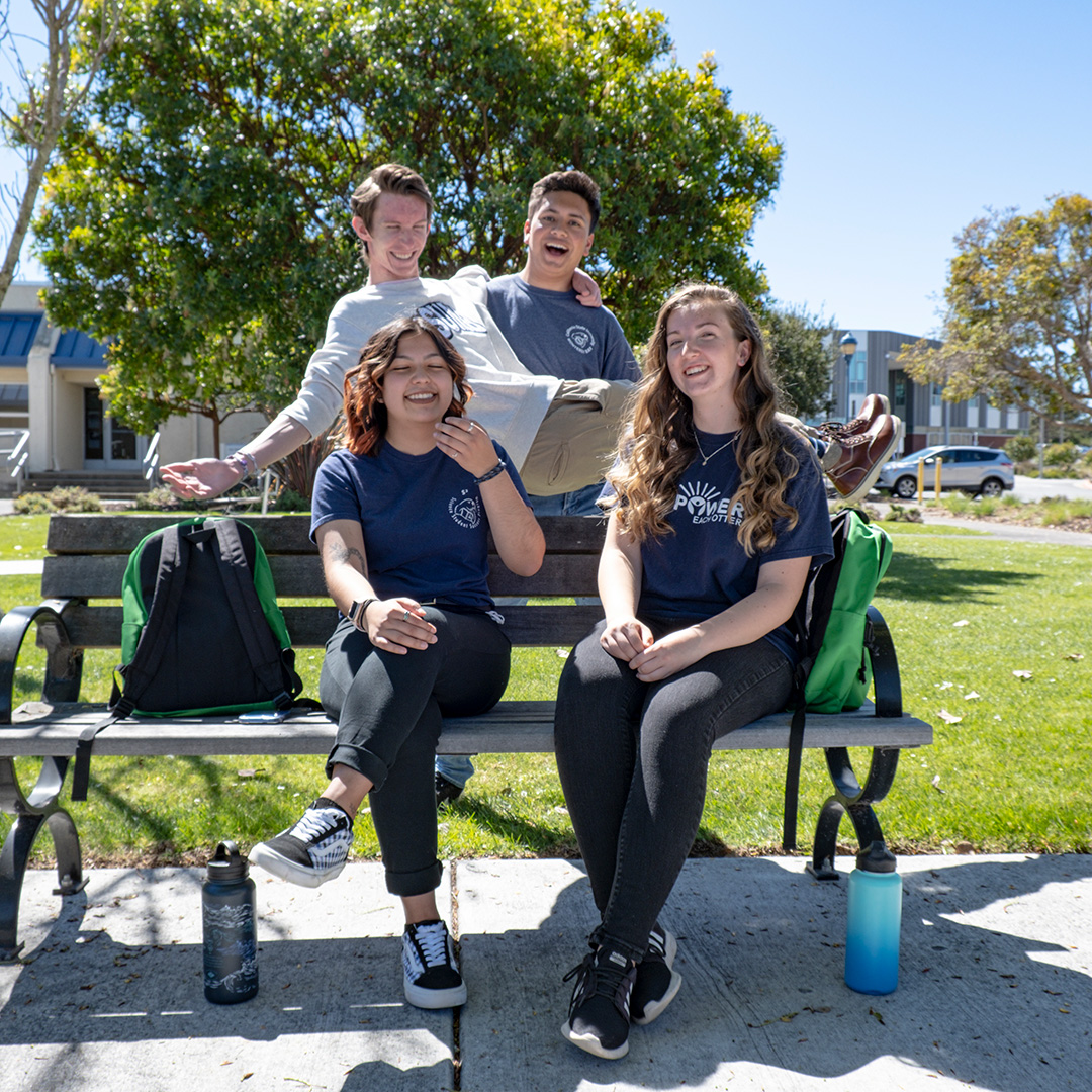 Four students wearing CSUMB gear sitting on a bench laughing.