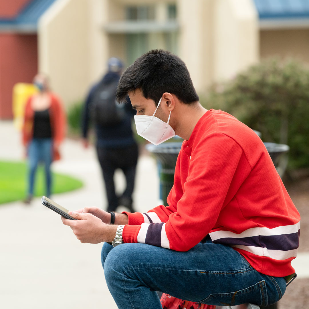 A student wears a mask while scrolling through his ipad outside.