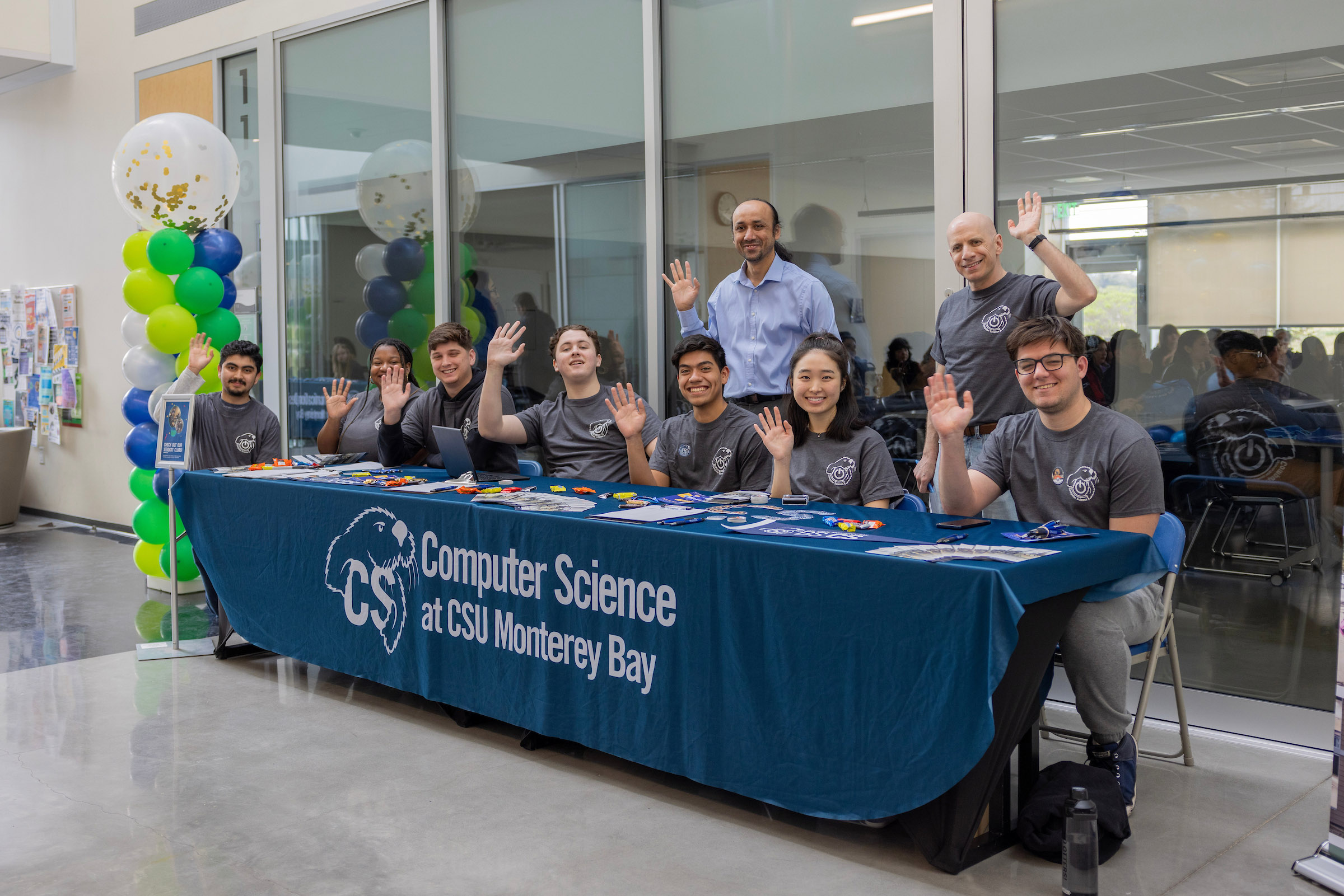 Students and staff waving behind a table that says Computer Science at CSU Monterey Bay.