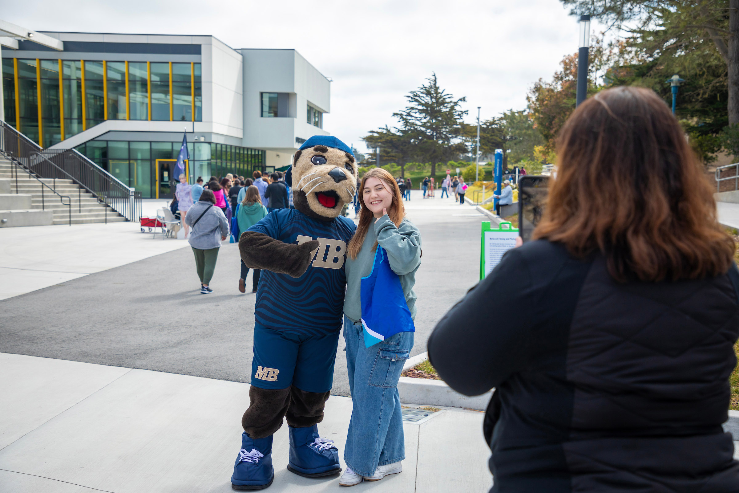 CSUMB sea otter mascot, Monte, stands next to a smiling, prospective student while a parent takes a cellphone photo.